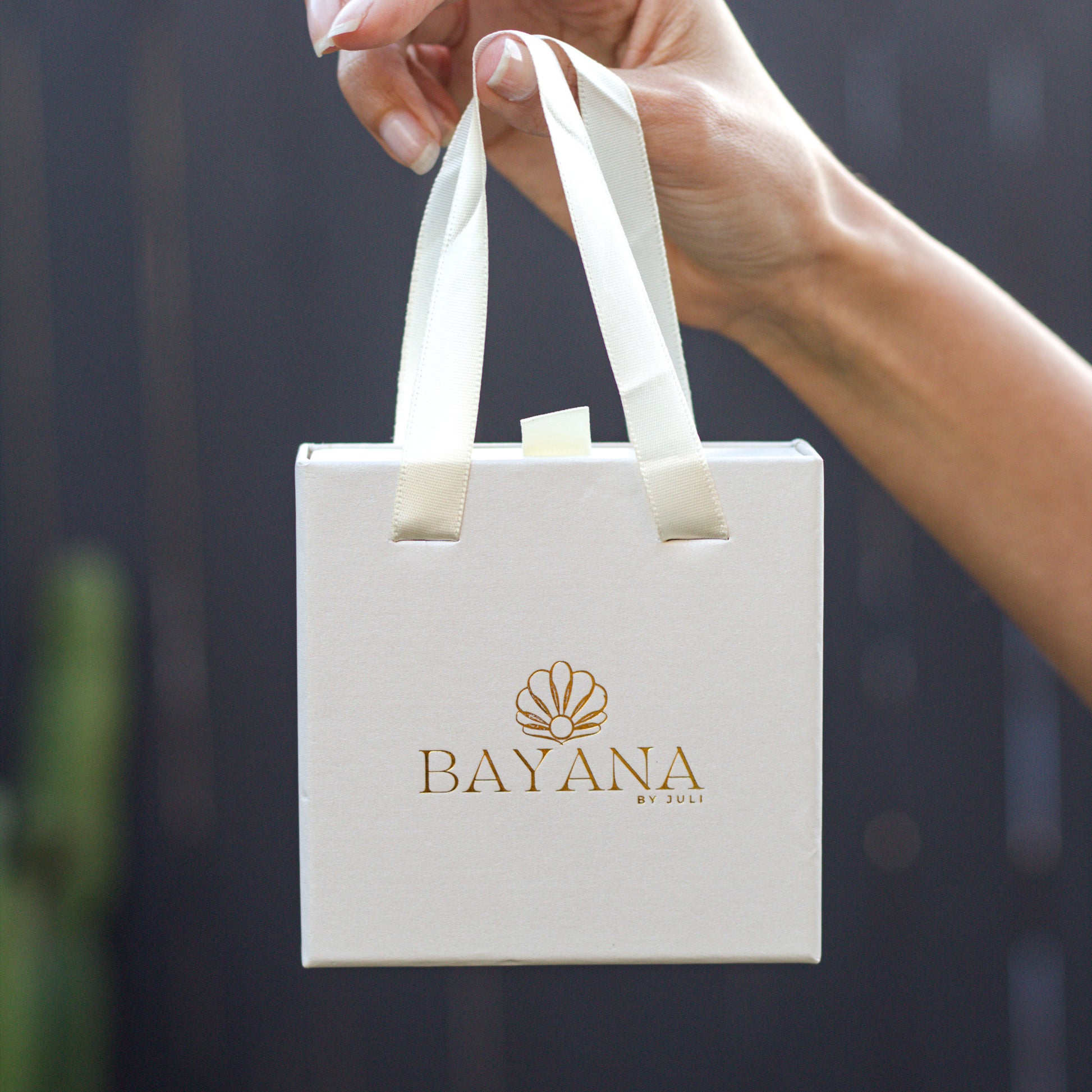 White gift bag with 'BAYANA' logo held by a hand against a dark background